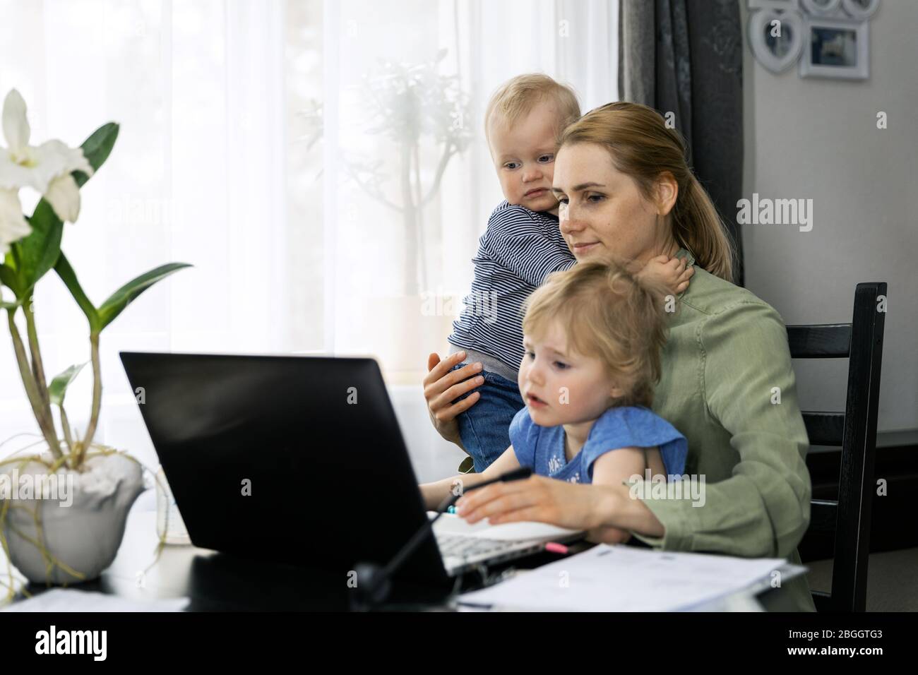 madre che lavora da casa. uso di un computer portatile alla scrivania con bambini in braccio Foto Stock