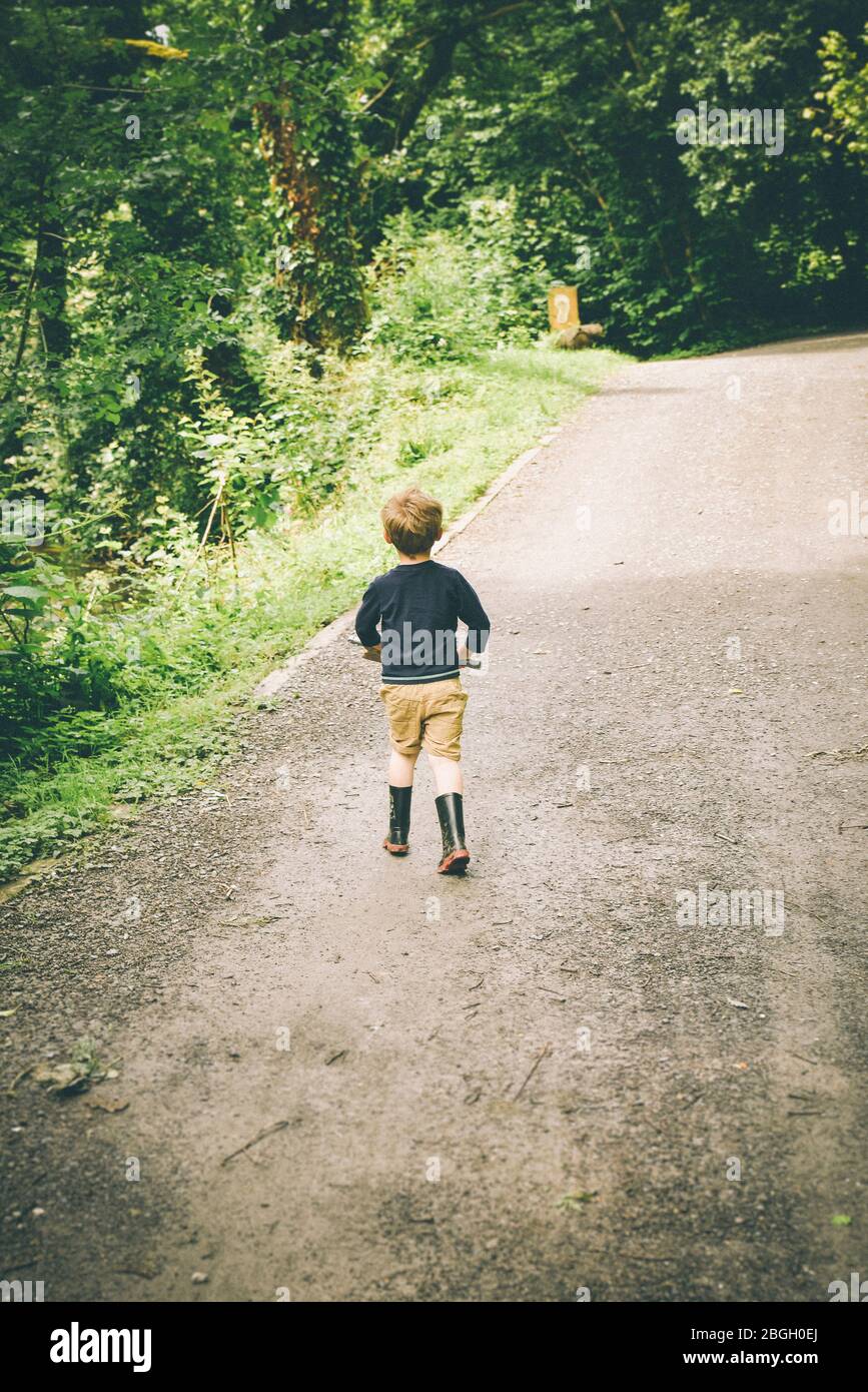 Vista posteriore del ragazzo giovane che cammina lungo la strada rurale in Shorts e stivali di gomma Foto Stock