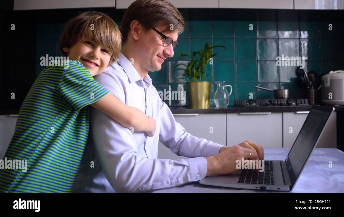 Un uomo lavora in cucina a un computer portatile con i suoi figli a casa. Padre con figlio. Telelavaggio a casa durante il periodo di quarantena del coronavirus Covid 19. Foto Stock