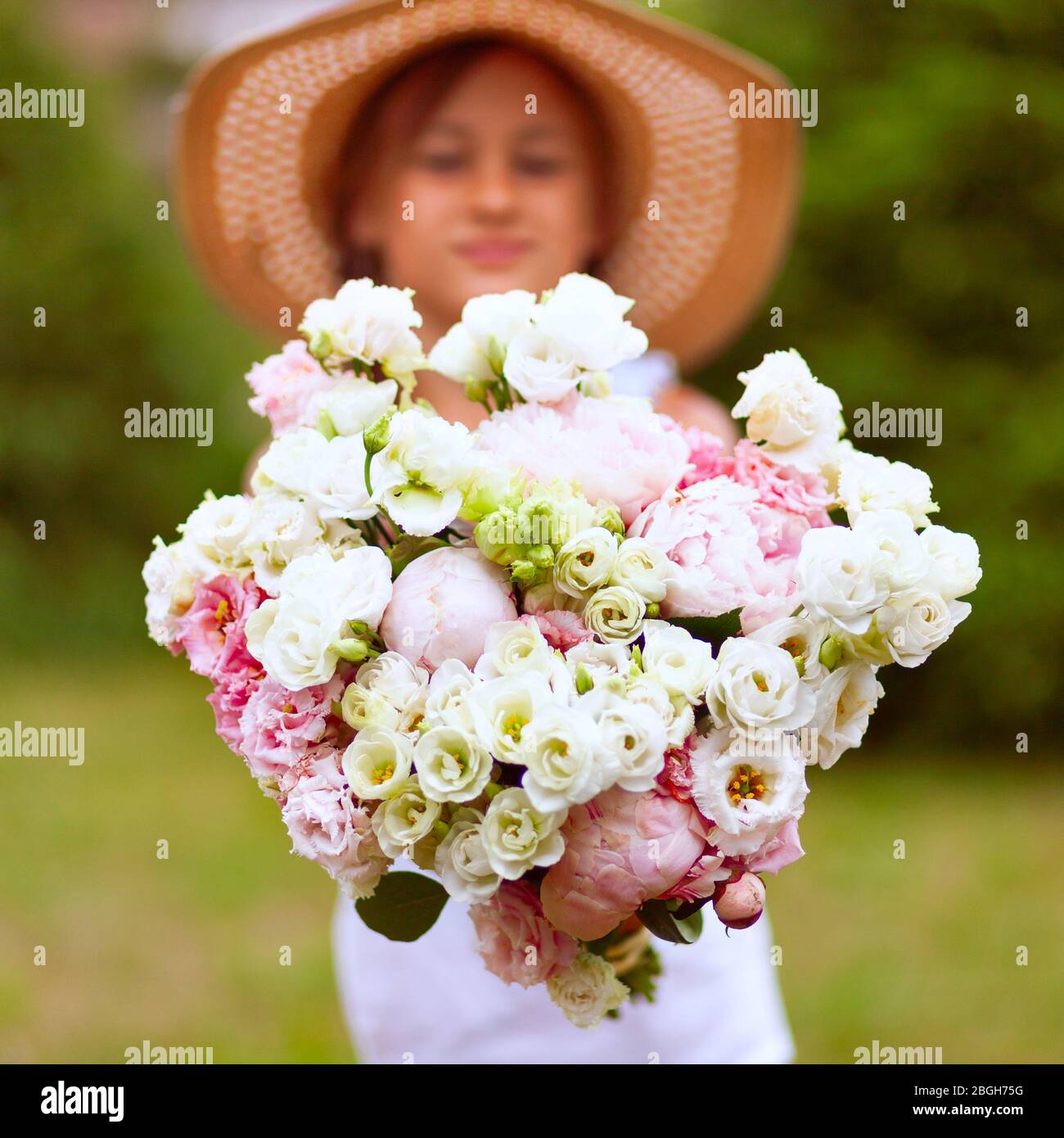 Un grande bouquet di peonie bianco-rosa nelle mani di una ragazza. Un bambino in un cappello di paglia con ampio bordo su uno sfondo di vegetazione verde. Giorno di sole. Foto Stock