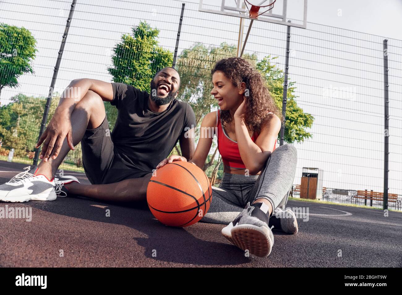Attività all'aperto. Coppia africana seduta con palla su campo di basket ridendo gioioso Foto Stock