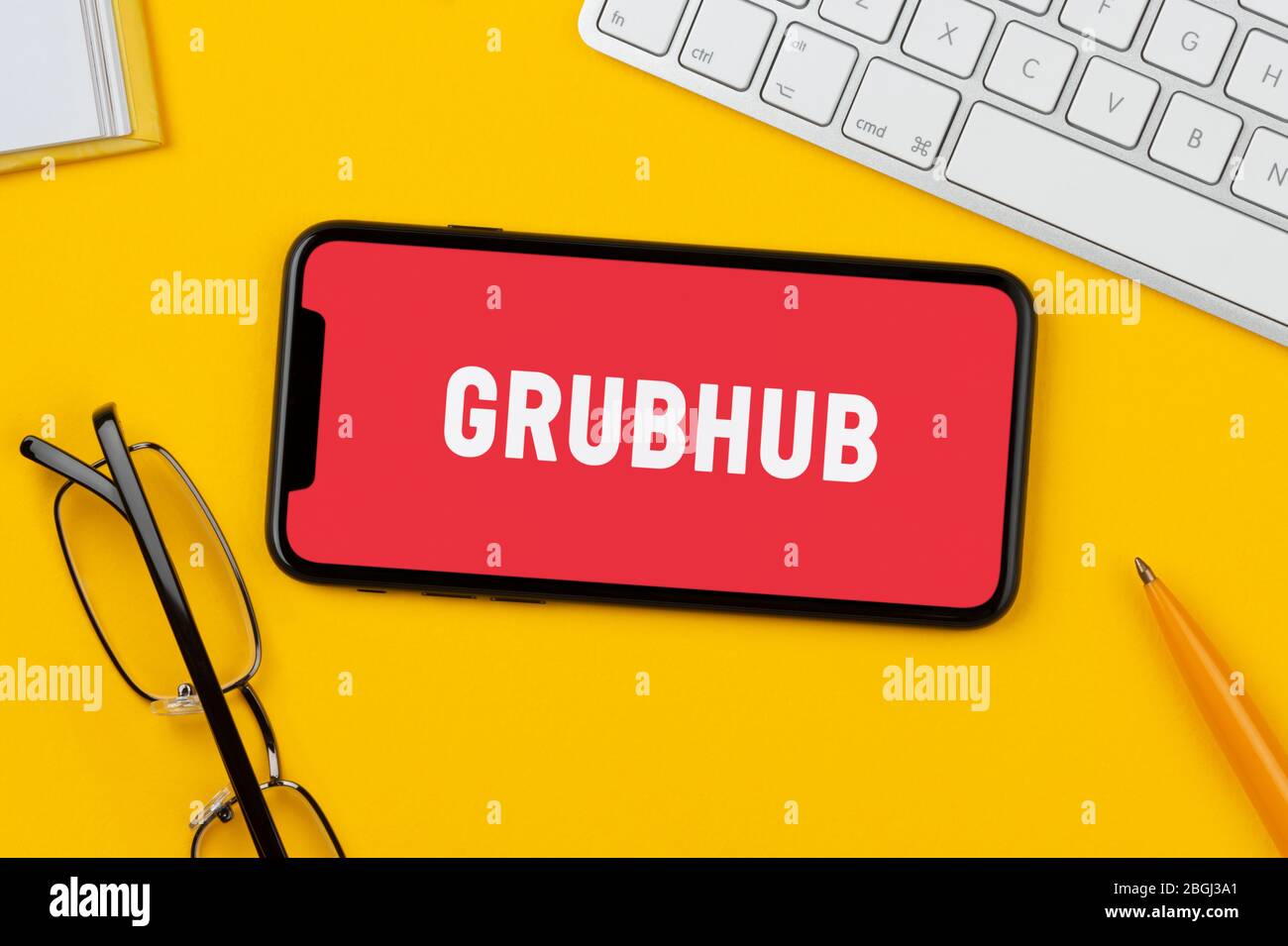 Uno smartphone con il logo Grubbuhub è posizionato su uno sfondo giallo insieme a tastiera, occhiali, penna e libro (solo per uso editoriale). Foto Stock