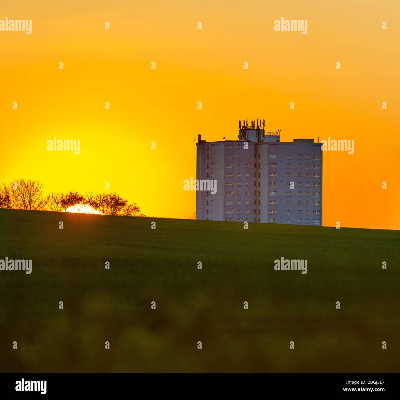 Immagine manipolata del sole che tramonta dietro un blocco di appartamenti a più piani con un campo verde in primo piano sotto un cielo arancione. Foto Stock