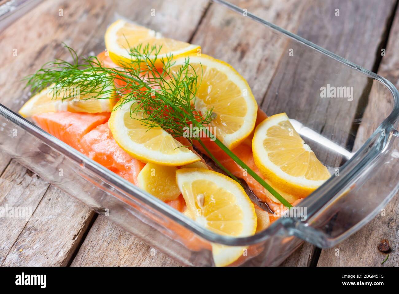 Filetti di pesce rosso o salmone in vetro da forno su sfondo di legno. Pesce fresco, limone e aneto per cucinare gli ingredienti. Foto Stock