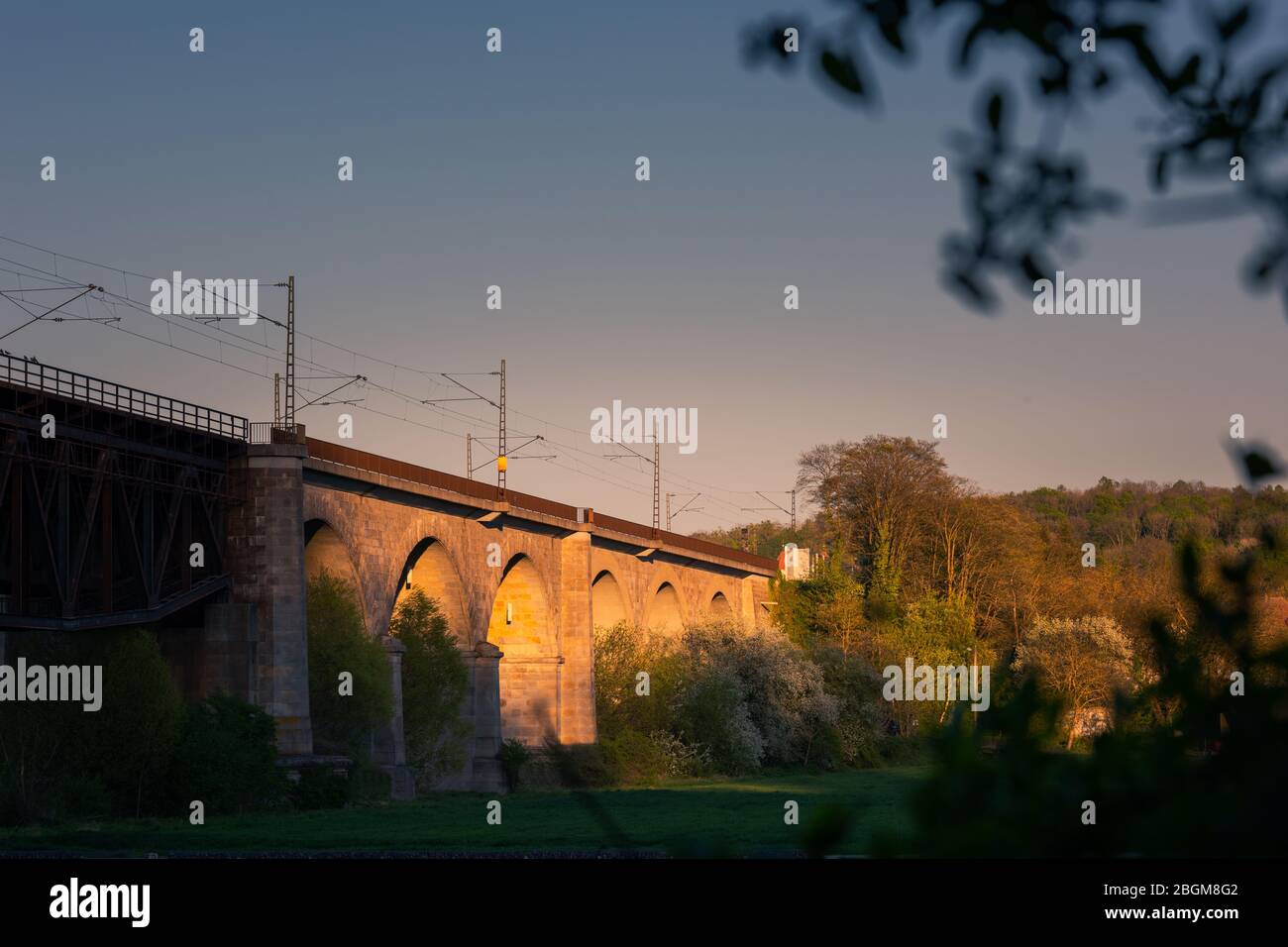 Ponte ferroviario di Mariaort con archi in pietra e linea di contatto aerea su campo verde che si illumina nel tardo pomeriggio luce del sole a Regensburg, Bava Foto Stock