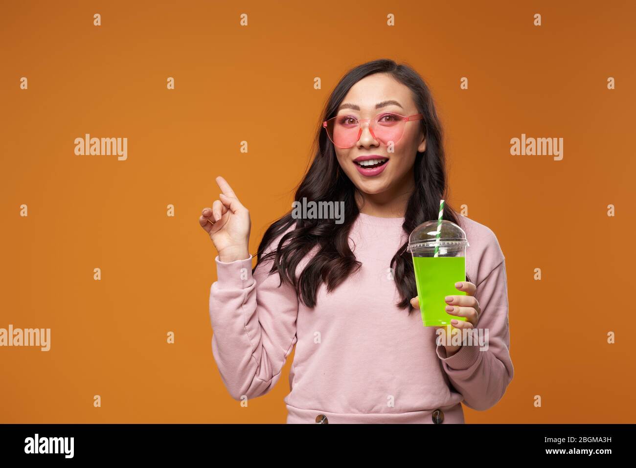 Donna asiatica in bicchieri rosa con cocktail, puntando con la mano a spazio vuoto su sfondo giallo vuoto in studio Foto Stock