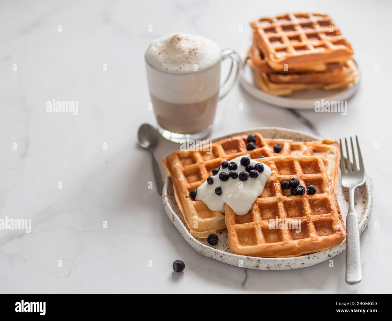 Deliziosi waffle belgi fatti in casa con yogurt greco, mirtilli e cappuccino su sfondo di marmo bianco. Colazione perfetta con spazio per testi o disegni Foto Stock