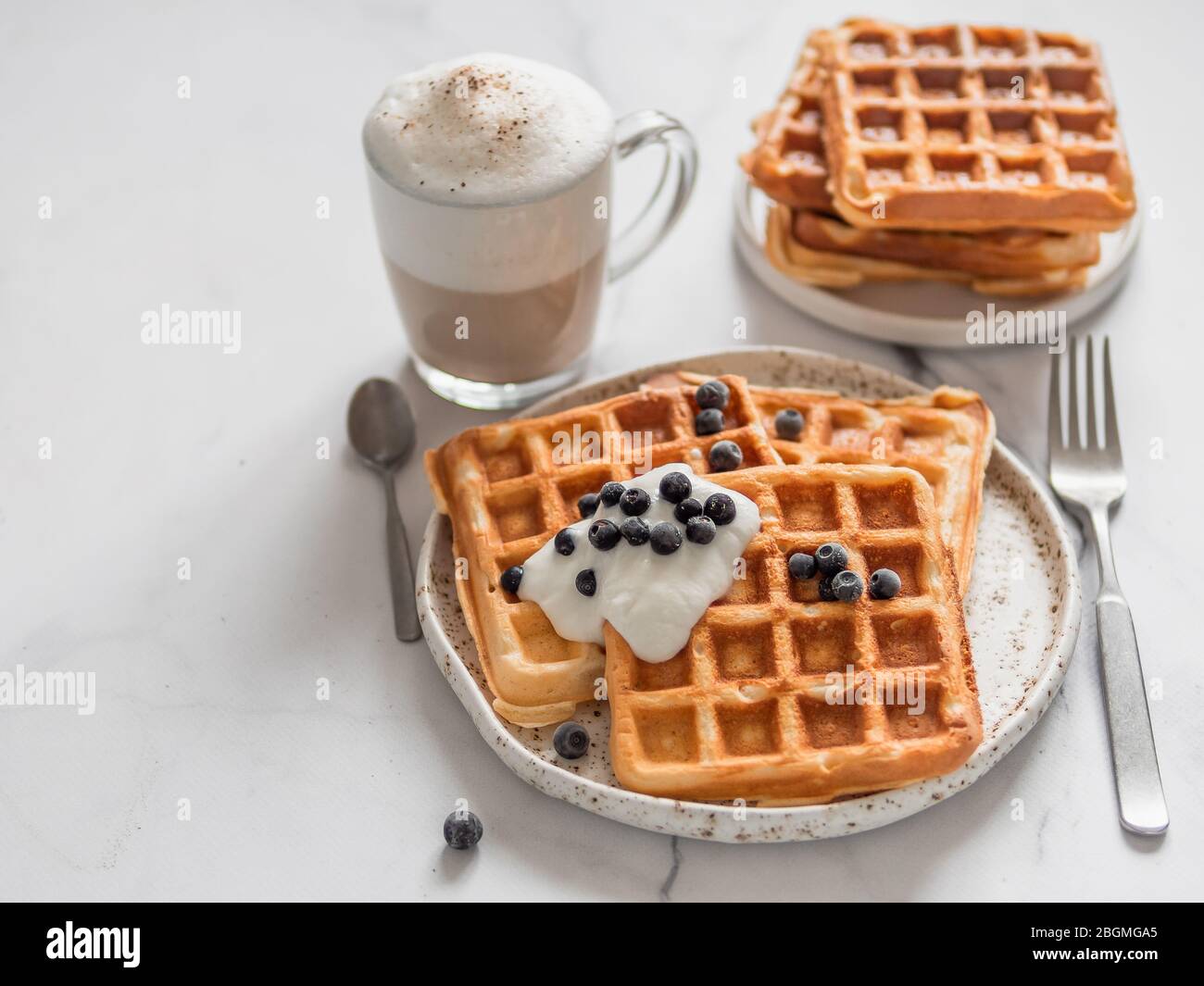 Deliziosi waffle belgi fatti in casa con yogurt greco, mirtilli e cappuccino su sfondo di marmo bianco. Colazione perfetta con spazio per testi o disegni Foto Stock