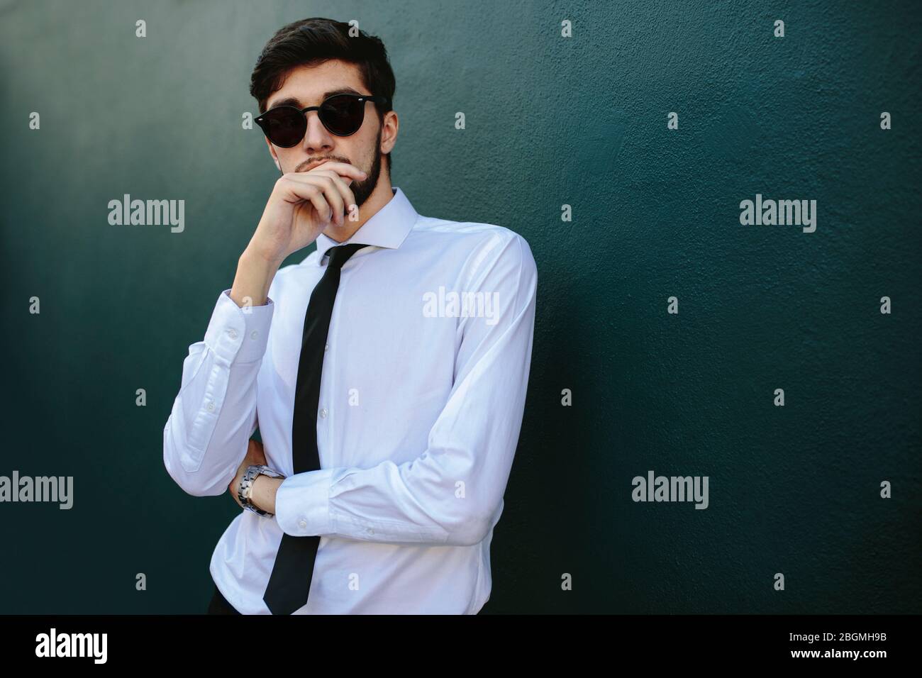 Giovane uomo d'affari vestito elegantemente in piedi da una parete scura. Uomo in formalwear posa in occhiali da sole. Foto Stock