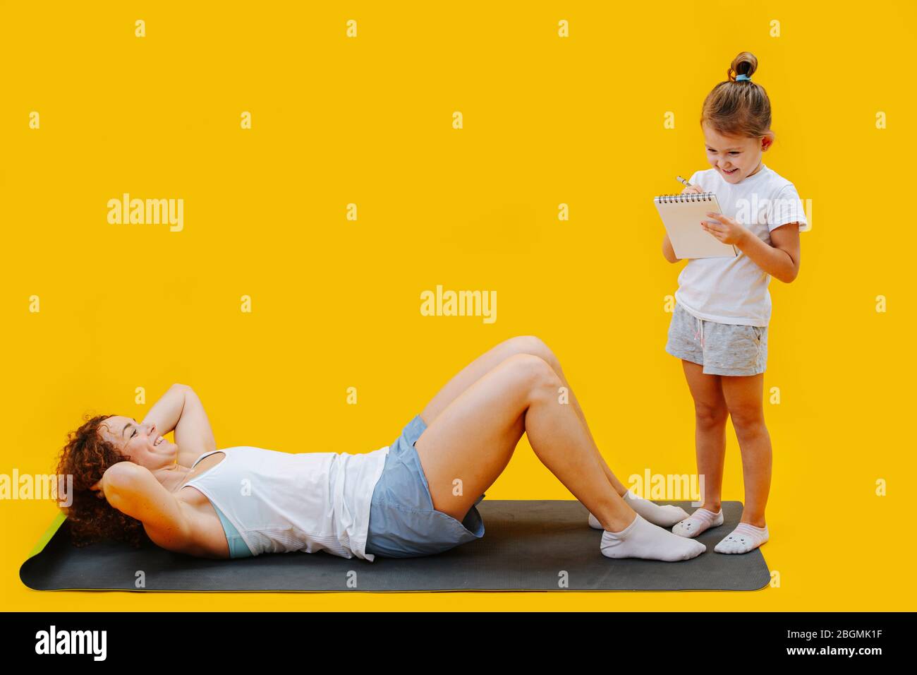 Figlia tenendo le linguette sugli esercizi fisici della mamma su sfondo giallo Foto Stock