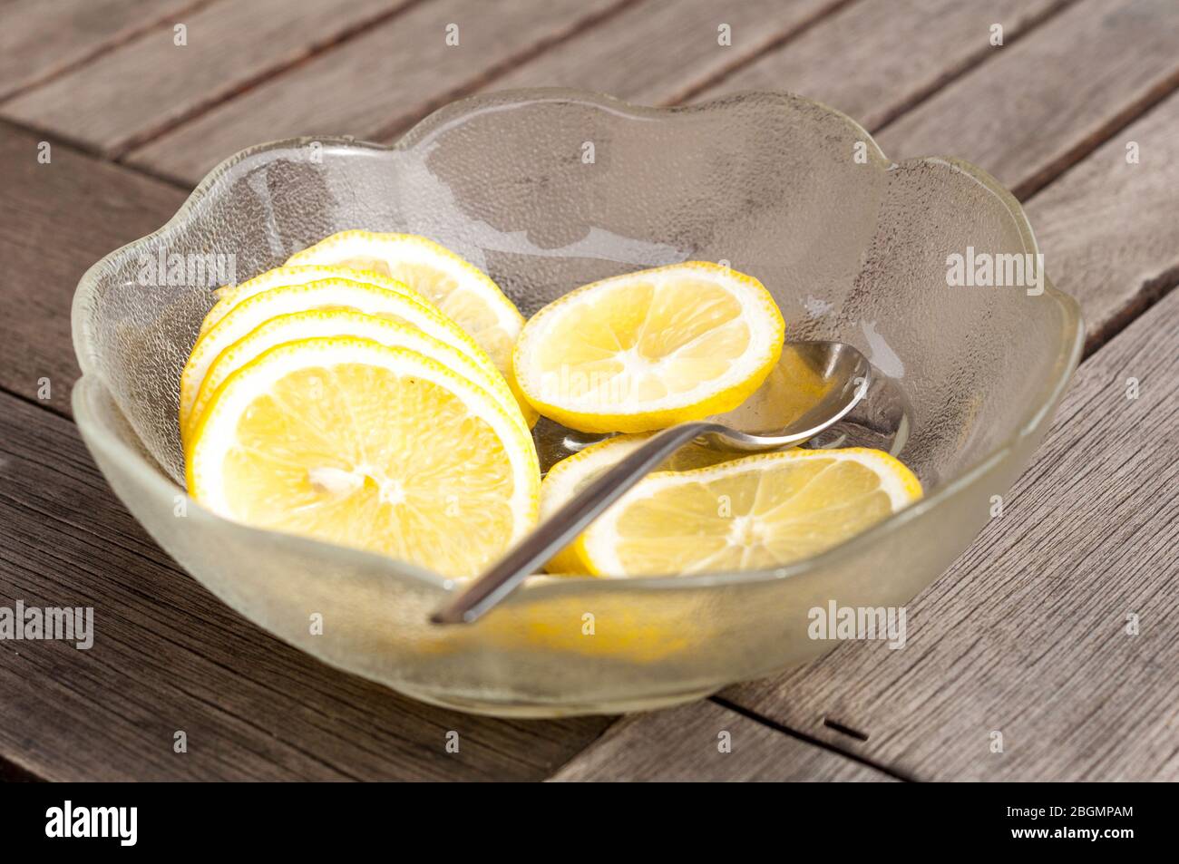 Una ciotola di vetro riempita di cunei di limone si trova su un tavolo di legno. Con il cucchiaio, le fette vengono poste in acqua rinfrescante o limonata. Foto Stock