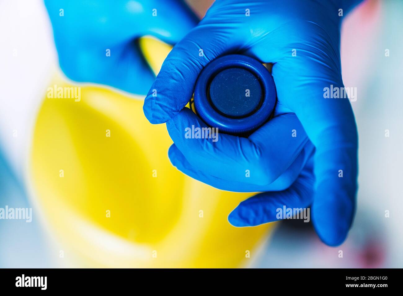 Mano con guanti in lattice blu che aprono una bottiglia di candeggina gialla. Foto Stock
