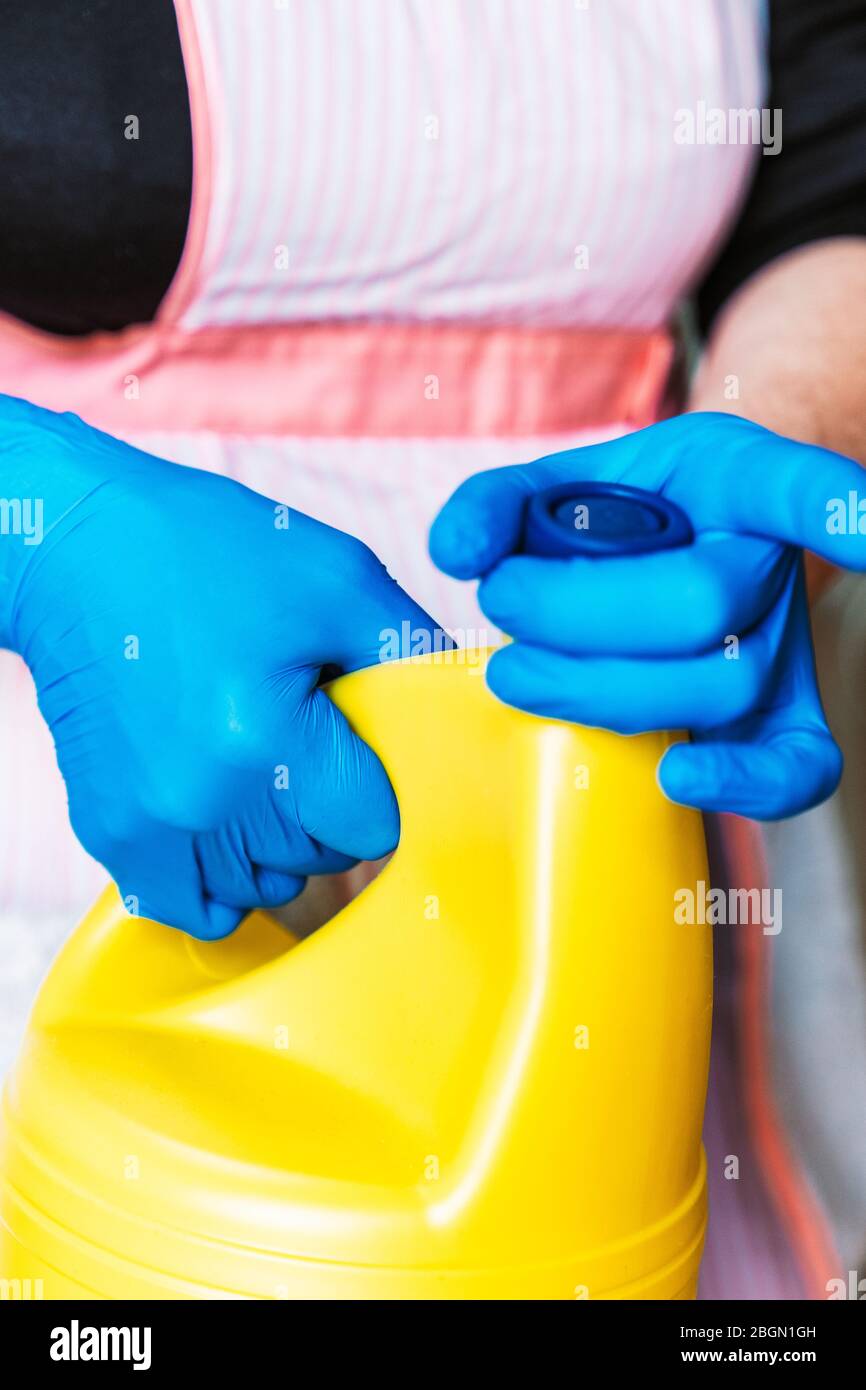 Donna con guanti in lattice blu e grembiule che apre una bottiglia di candeggina gialla. Foto Stock