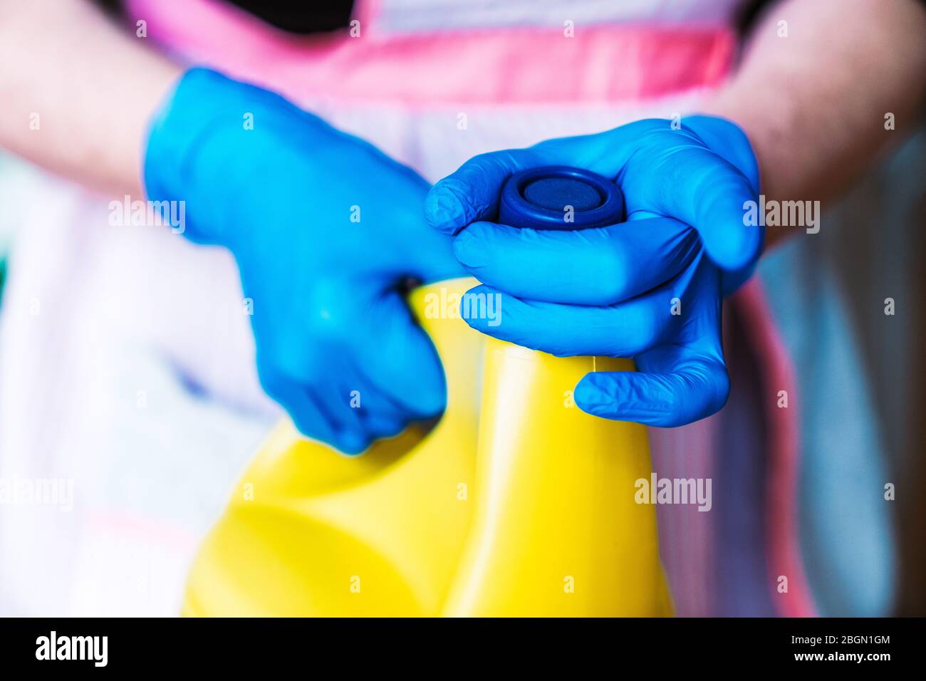 Casalinga con guanti in lattice blu e grembiule che apre una bottiglia gialla di candeggina. Foto Stock