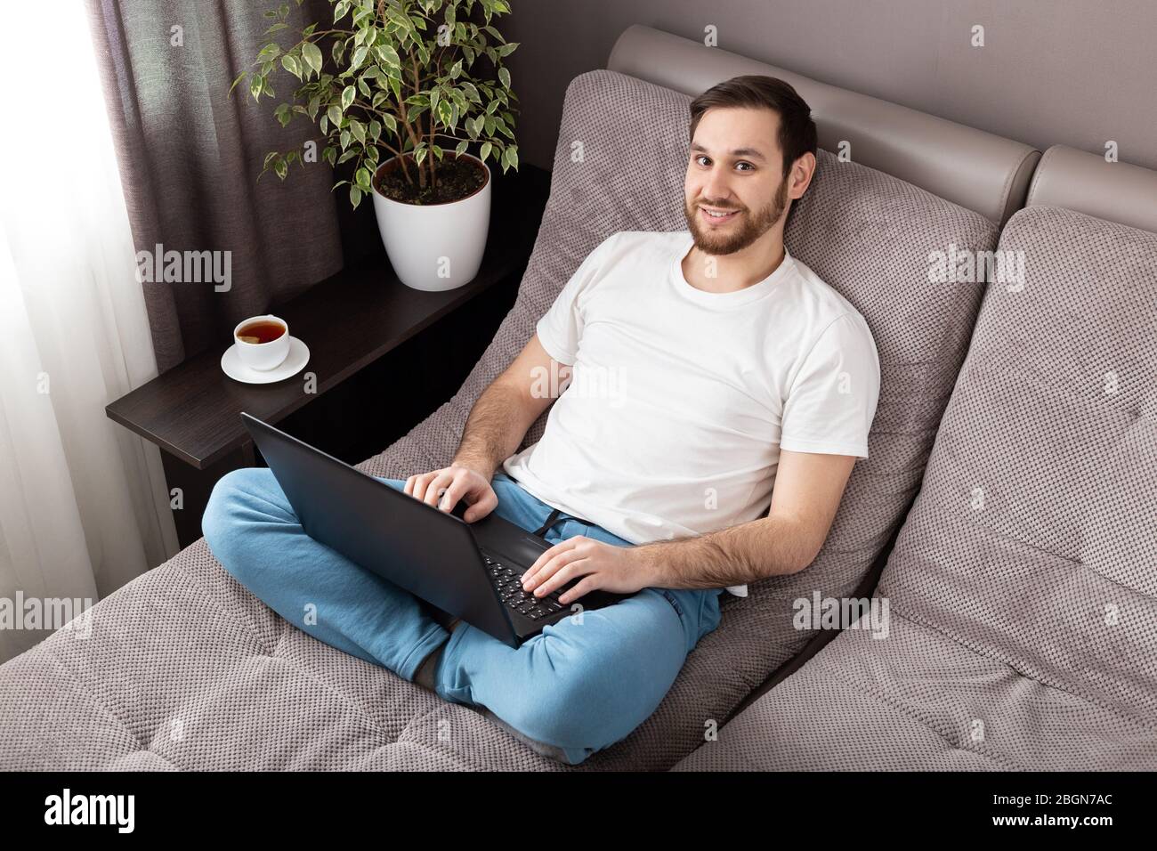 Luogo di lavoro domestico. Sorridente felice uomo freelancer che lavora da casa utilizzando il laptop. Comodo ufficio sul divano. Coronavirus covid-19 sociale che si distanziano Foto Stock
