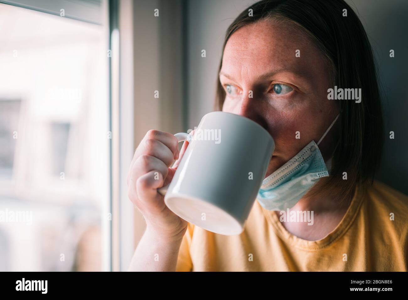 Donna in auto-isolamento durante l'epidemia di virus bere caffè e guardare attraverso la finestra. Donna preoccupata con maschera chirurgica protettiva. Foto Stock