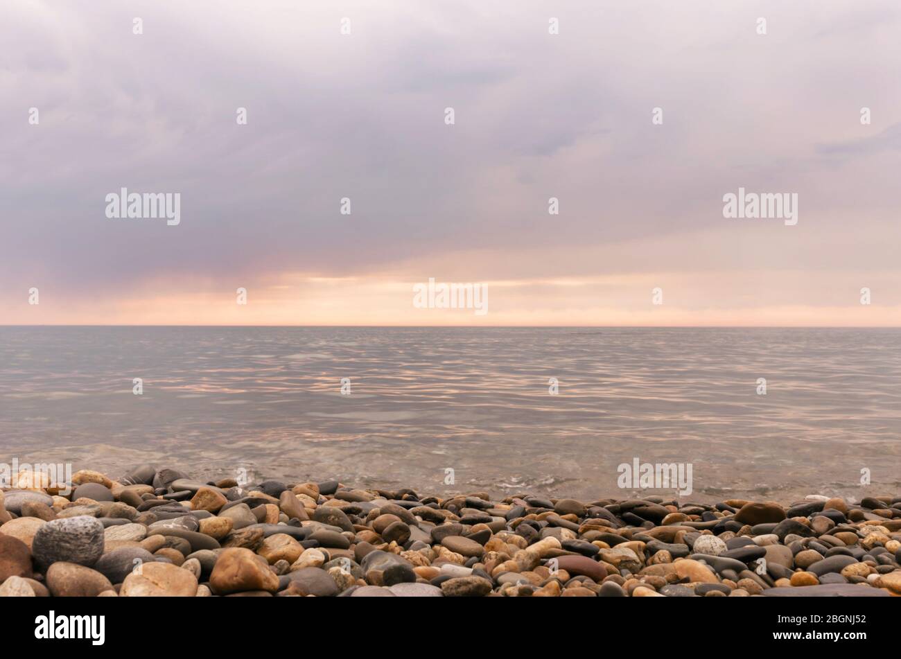 Pioggia nuvole sopra il mare al tramonto. I ciottoli sulla spiaggia di close-up, poco onde sull'acqua e nuvole colorate sfumature di rosa. Foto Stock