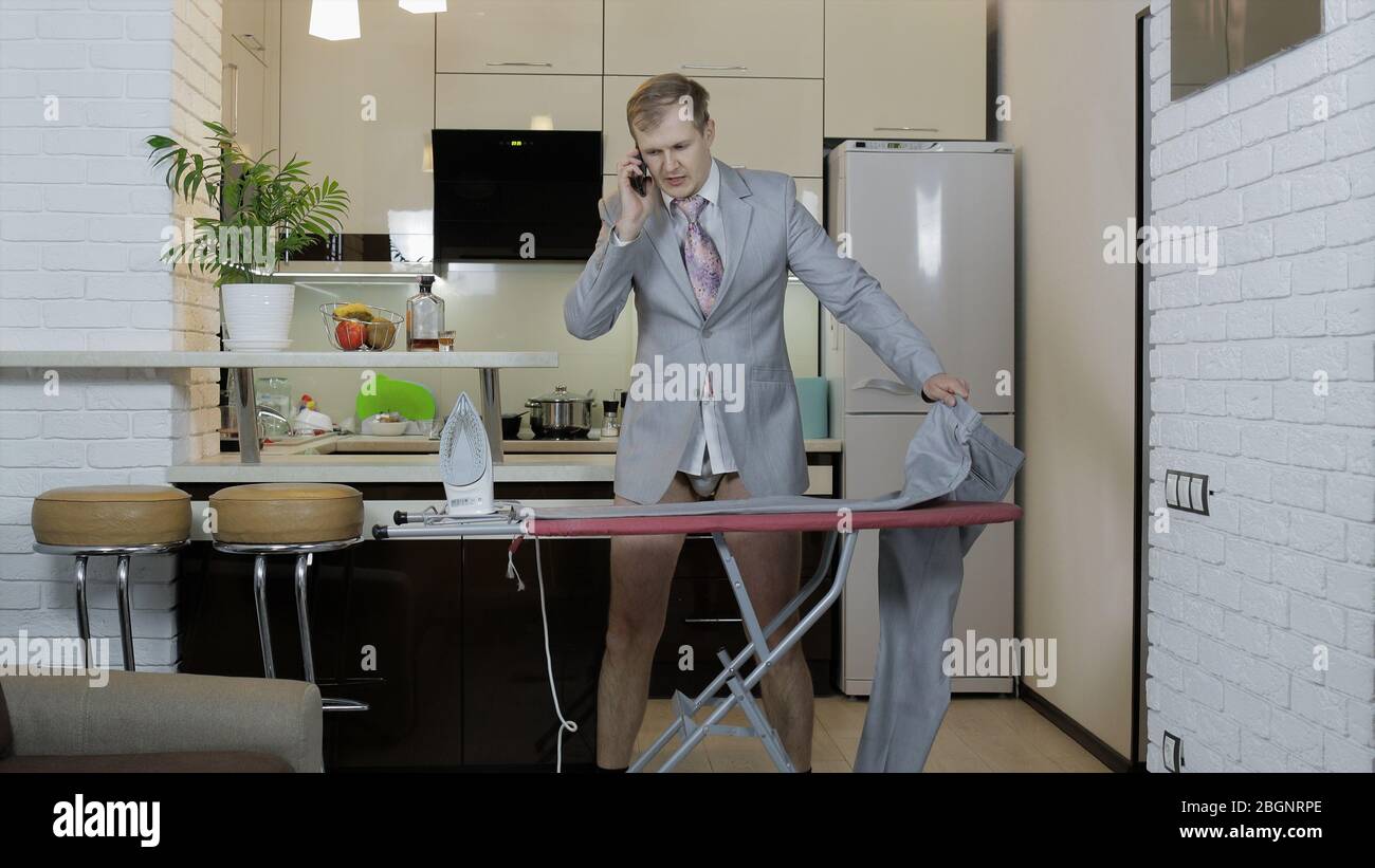 bell'uomo con cravatta, camicia bianca e pantaloni da stiro mentre parla al telefono. in piedi vicino alla cucina di casa. uomo d'affari a casa che si prepara per una riunione d'affari. stira i suoi vestiti Foto Stock