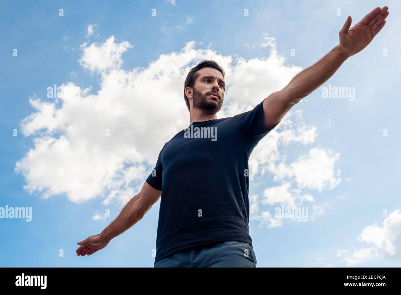 Un uomo che indossa una camicia nera e pantaloncini verdi scuri allunga le braccia in una posa guerriero sperando di correggere il suo corpo davanti a un blu brillante s. Foto Stock