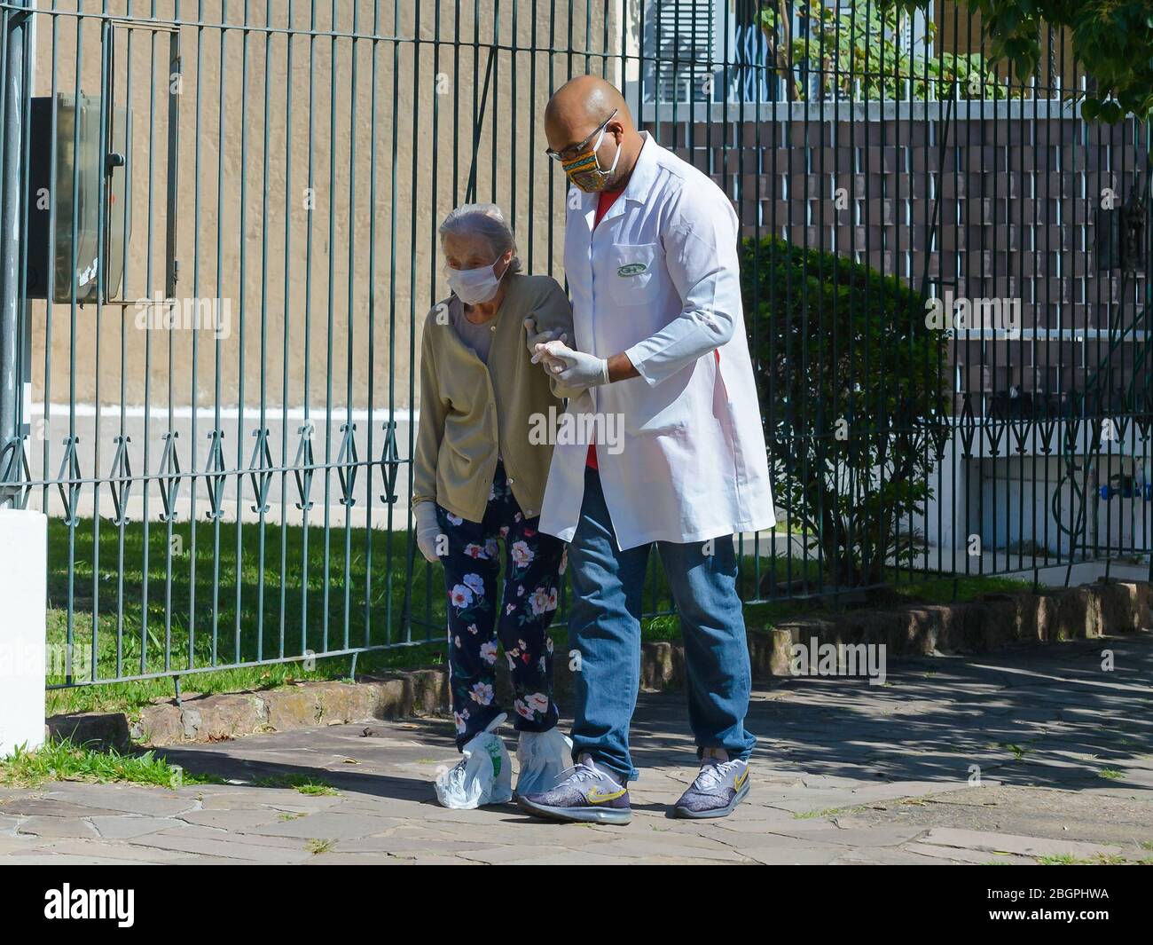 Donna anziana che cammina all'aperto con l'aiuto di un operatore sanitario che utilizza maschere facciali e guanti durante l'epidemia di Coronavirus. Operatore sanitario in Brasile. Foto Stock