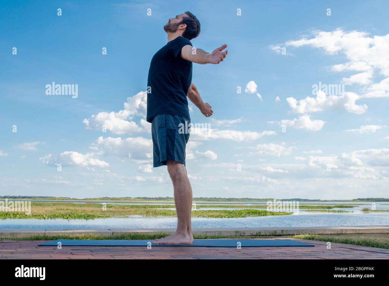 Uomo che indossa una camicia nera e pantaloncini verdi scuri si alza dritto con le braccia aperte, correggendo la posizione del corpo davanti a un blu brillante Foto Stock