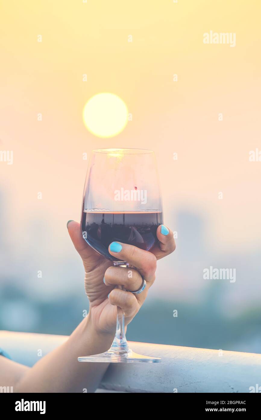 Mano della donna che tiene i bicchieri di vino rosso sul balcone durante il tramonto, concetto di festa Foto Stock