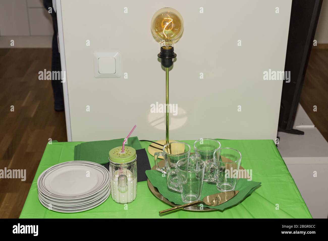 Vista ravvicinata di un grazioso tavolino con piatti bianchi e bicchieri vuoti preparati per la festa di compleanno di casa. Lampada da tavolo moderna su retro di tela verde Foto Stock