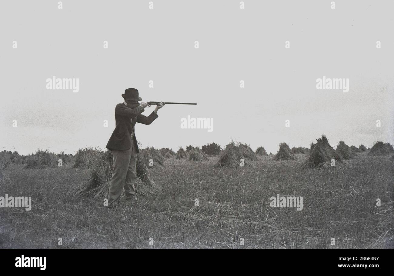 Anni '1930, storico agricoltore gentiluomo che indossa una giacca sportiva e un cappello in piedi in un campo di fieno falciato con piccoli fieno, con il suo fucile tenuto in alto mirato a un bersaglio, forse un coniglio, Devon, Inghilterra, Regno Unito. Foto Stock
