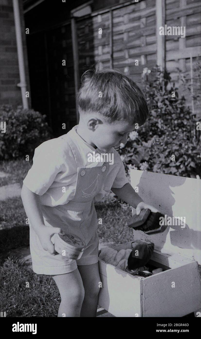 1962, storico, estivo e un bambino che indossa salopette corte, all'aperto che gioca con la "scatola per scarpe" di suo padre, una scatola di legno fatta a mano con spazzole, panni e lucidatura per scarpe, Inghilterra, Regno Unito. Foto Stock