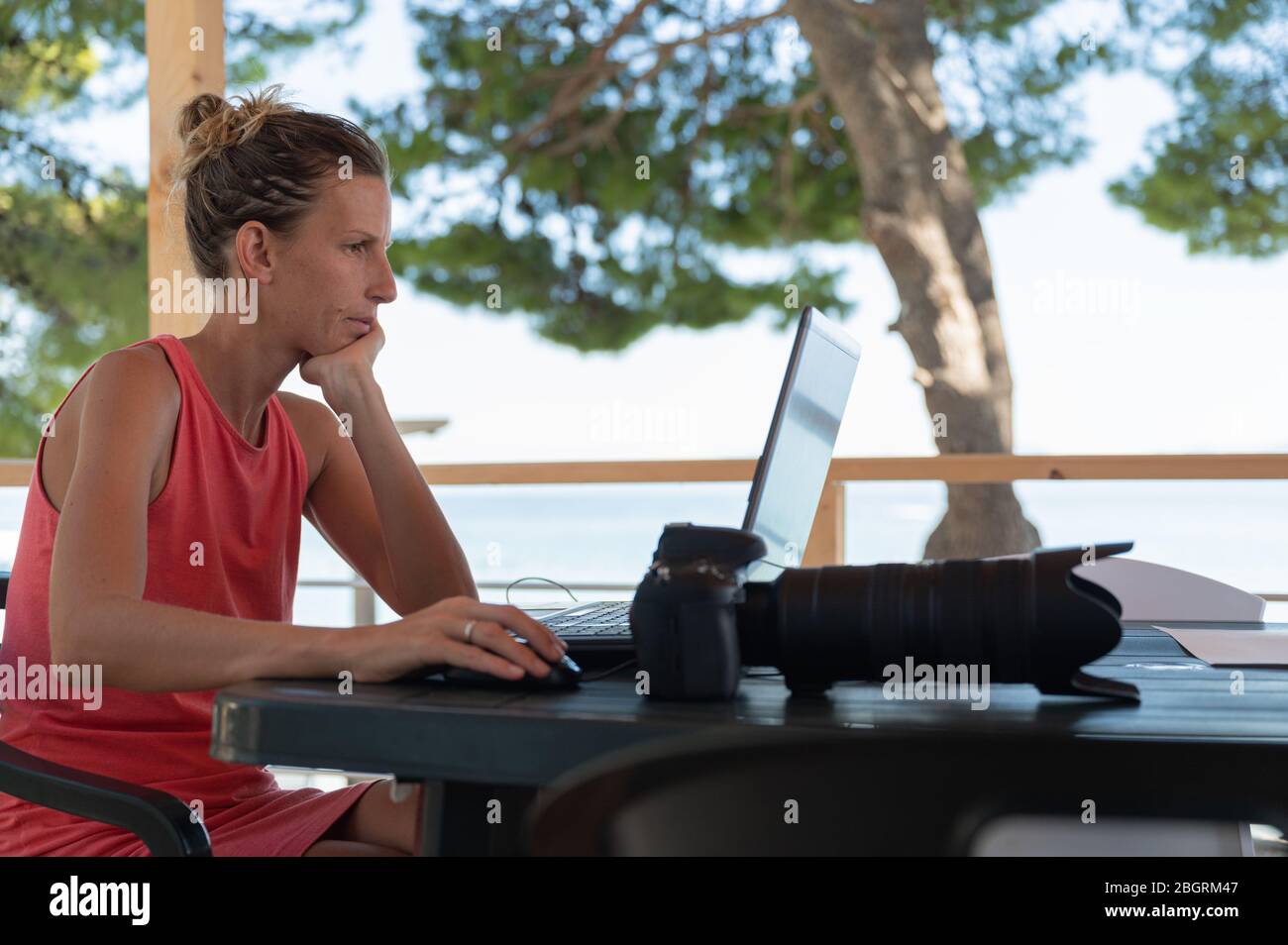 Giovane fotografo femminile che controlla e modifica le foto sul suo computer portatile, seduto a una scrivania all'aperto nel resort estivo. Foto Stock