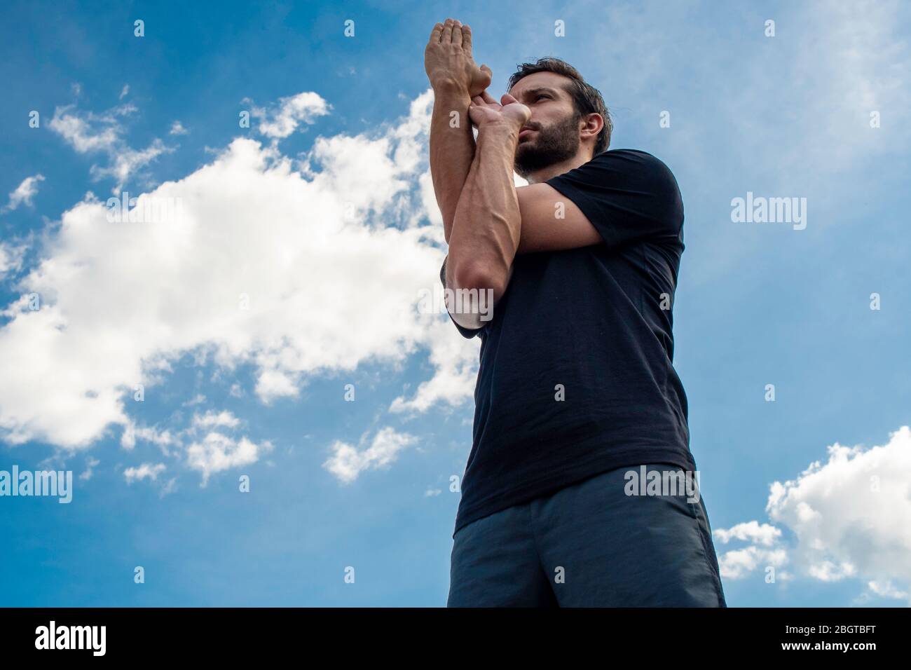 Uomo che indossa una camicia nera e pantaloncini verdi scuri si alza dritto con le mani in posa aquila correggendo la posizione del corpo davanti a un blu brillante Foto Stock