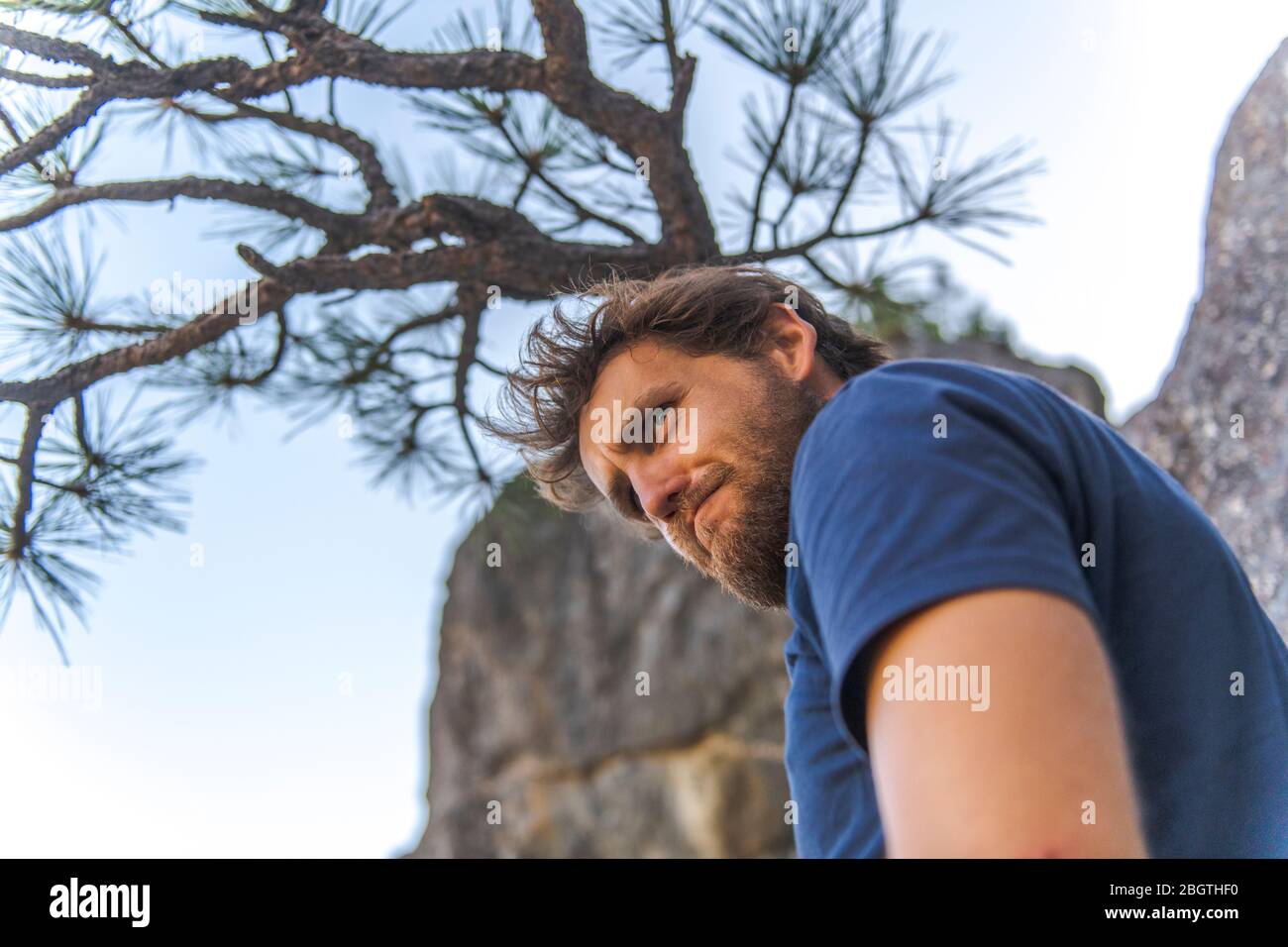 Uomo in camicia con barba che fa divertente faccia guardando sotto l'albero Foto Stock