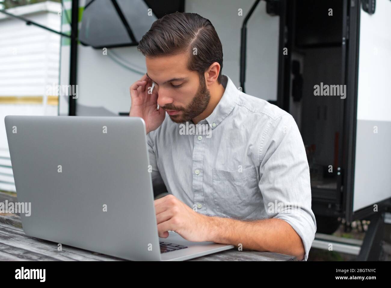 Un uomo che indossa una camicia da ufficio con bottoni grigi a manica lunga con pettine marrone affettato sui capelli che lavora su un laptop su un tavolo da picnic a distanza vicino alla sua ca Foto Stock