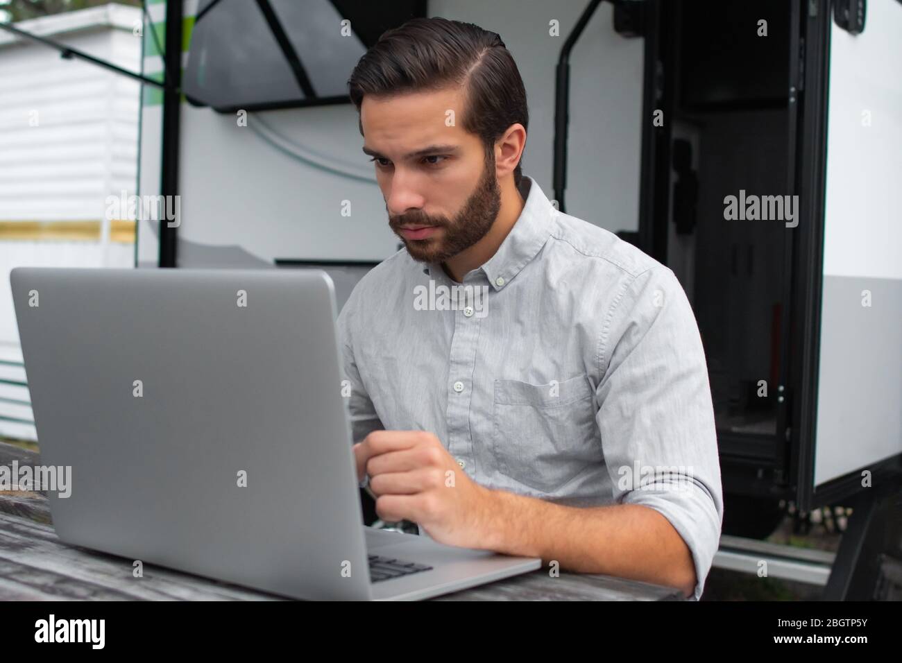 Un uomo che indossa una camicia da ufficio con bottoni grigi a manica lunga con pettine marrone affettato sui capelli che lavora su un laptop su un tavolo da picnic a distanza vicino alla sua ca Foto Stock