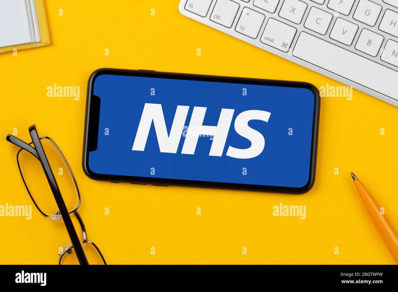 Uno smartphone con il logo NHS è posizionato su uno sfondo giallo insieme a tastiera, occhiali, penna e libro (solo per uso editoriale). Foto Stock