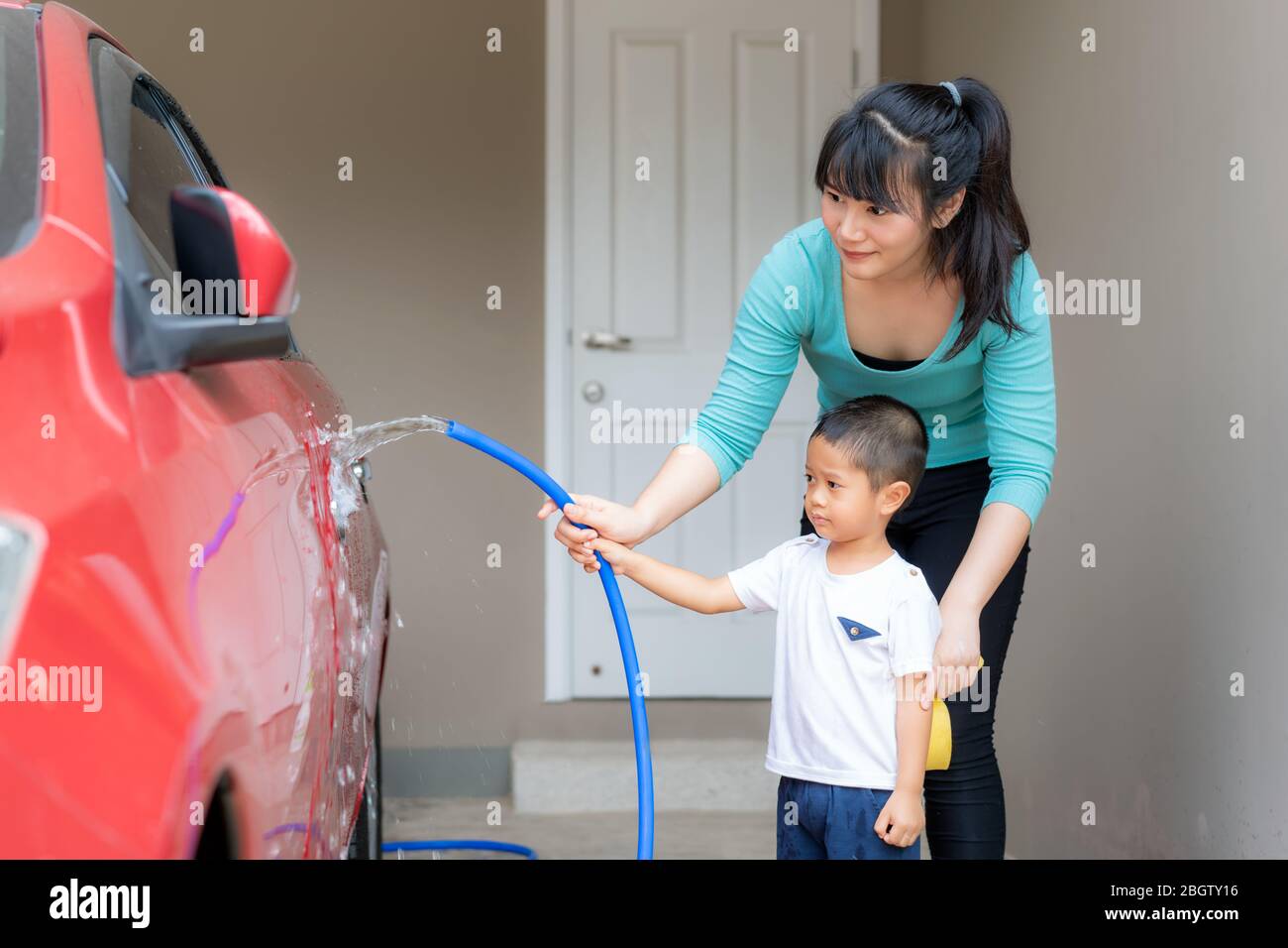 Madre asiatica e suo figlio che ha felice in casa lavatrice proprietario auto rossa utilizzando tubo spruzzi d'acqua durante il soggiorno a casa utilizzando il tempo libero circa la loro coda Foto Stock
