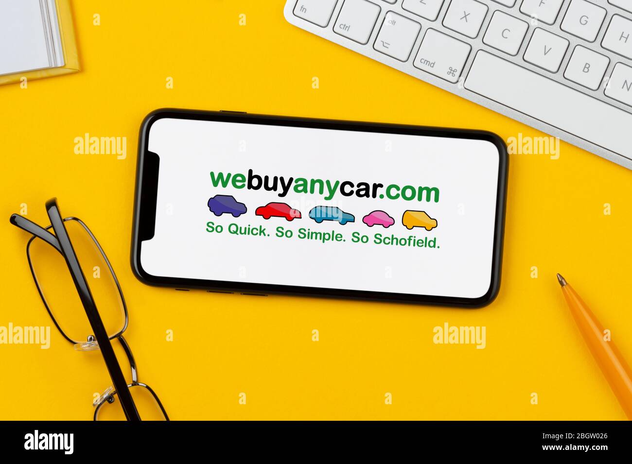 Uno smartphone con il logo webuyanycar è posizionato su uno sfondo giallo insieme a tastiera, occhiali, penna e libro (solo per uso editoriale). Foto Stock