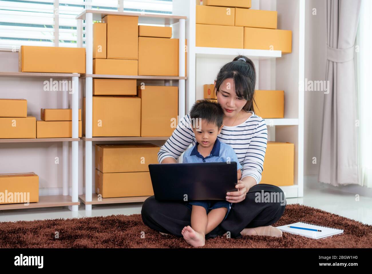 La donna d'affari del proprietario asiatico lavora a casa per fare shopping online, chiacchiera con il cliente con il laptop mentre lei tiene suo figlio a casa. Concetto di mamma singola. Foto Stock