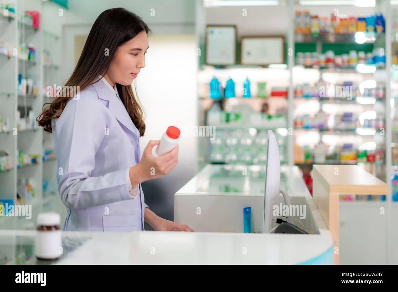 Fiducioso asiatici femmina giovane farmacista con sorriso tenendo una bottiglia di medicina e ricerca di quel prodotto nel database di computer in farmacia farmaci Foto Stock