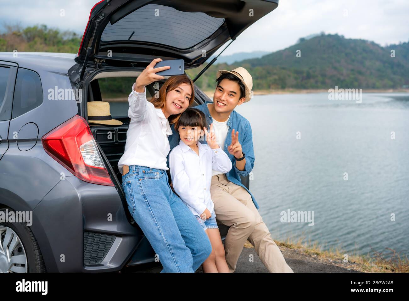 Ritratto della famiglia asiatica seduto in auto con padre, madre e figlia selfie con vista lago e montagna da smartphone, mentre la vacanza insieme in h. Foto Stock