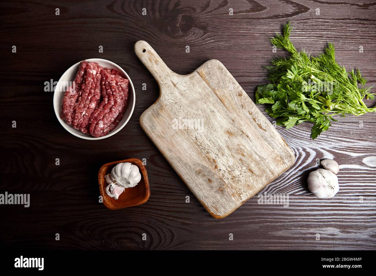 Cottura di carne macinata. Tagliere con condimenti freschi su un tavolo di legno, piatto. Teste e spicchi d'aglio bianchi, foglie di aneto verde e prezzemolo. Bo Foto Stock