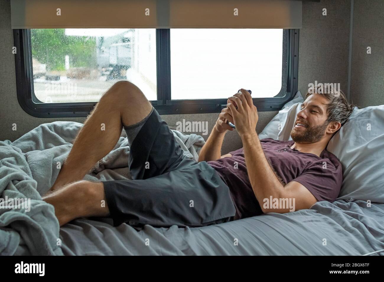 Un uomo ipster bearded bruno con capelli che indossa una t-shirt rossa si stende ridendo rilassandosi sul suo letto nel suo camper che parla con qualcuno sul suo smartphone su Foto Stock