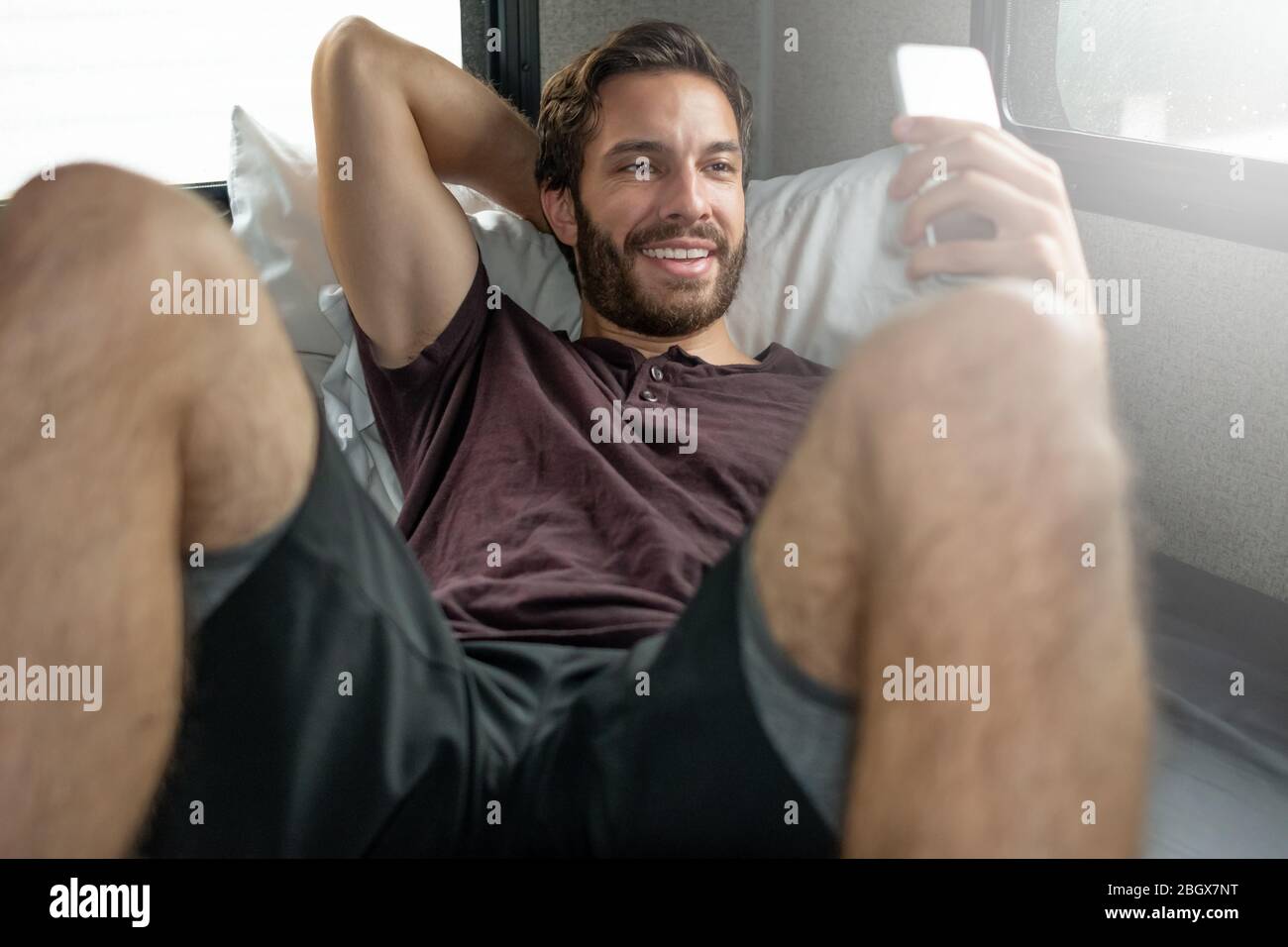 Un uomo ipster bearded bruno con capelli che indossa una t-shirt rossa si distende rilassandosi sul suo letto nel suo camper che parla con qualcuno sul suo smartphone su un nuvoloso Foto Stock