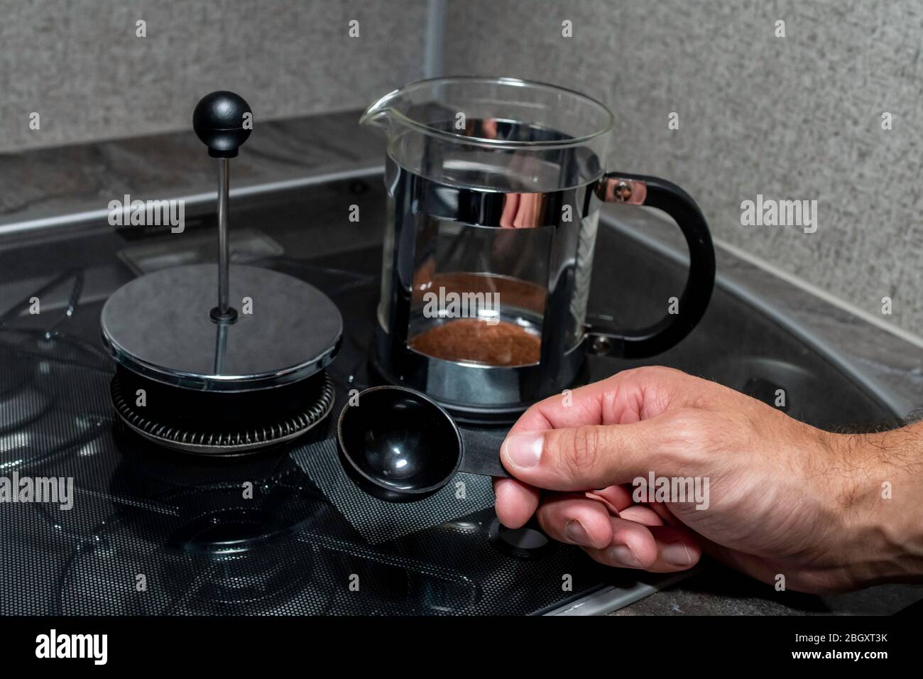 Primo piano sopra l'angolo della spalla di una persona mani di fissaggio caffè in una pressa francese di metallo nero e acciaio inossidabile su una piccola stufa coperta in gla Foto Stock