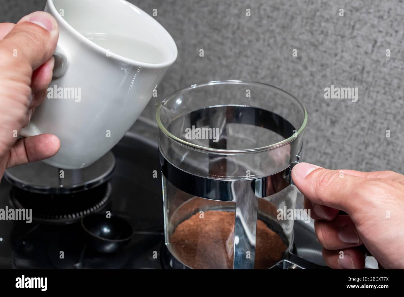 Primo piano sopra l'angolo della spalla di una persona mani di fissaggio caffè in una pressa francese di metallo nero e acciaio inossidabile su una piccola stufa coperta in gla Foto Stock