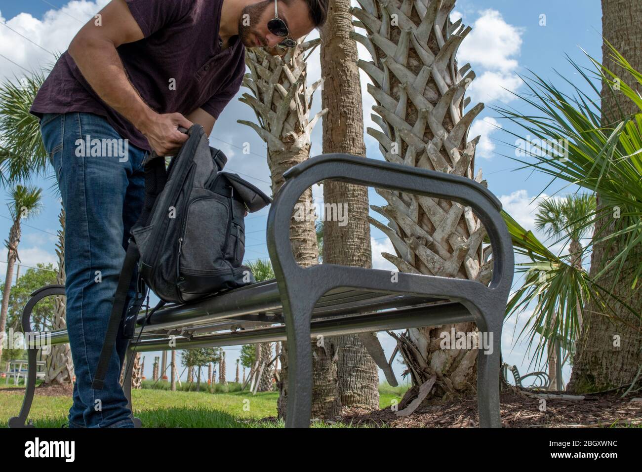 Un uomo che indossa una camicia rossa e jeans blu allunga il braccio verso l'esterno raggiungendo giù in uno zaino nero alla ricerca in una giornata di sole brillante su una panca parco dentro Foto Stock