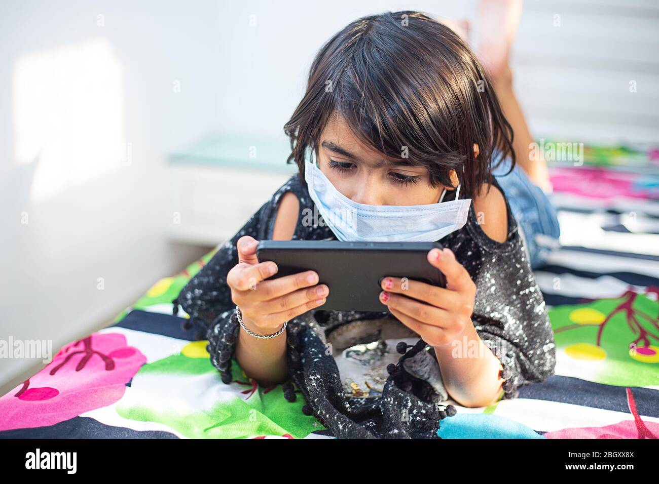 Giovane ragazza prescolare che indossa la maschera sdraiata su un letto in una posa rilassata, giocando al suo smartphone. Rimanere a casa durante la quarantena. Distan sociale Foto Stock