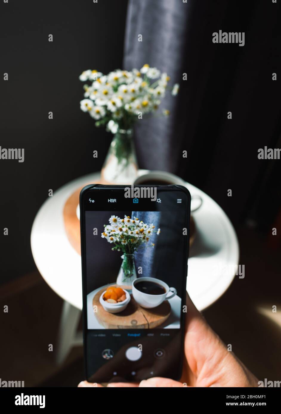 Mano con lo smartphone che fotografa una tazza di caffè con i fiori a casa. Foto Stock