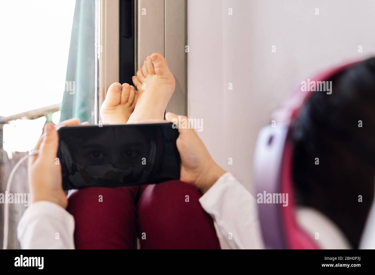 bambino seduto a casa con una cuffia e maschera protettiva riflessa sullo schermo del suo telefono cellulare, concetto di coronavirus e comunicazione in quarantena Foto Stock
