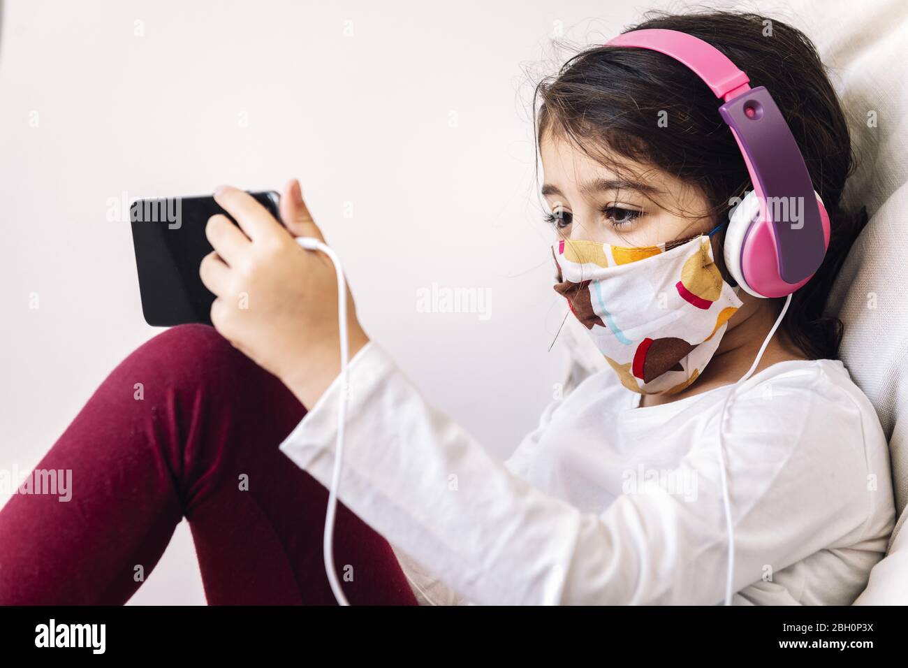 bambino seduto a casa con una cuffia e maschera protettiva facendo una videochiamata con il telefono, il concetto di coronavirus e la comunicazione in quarantena Foto Stock